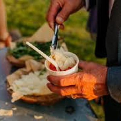 Een hand die een beker pasta vasthoudt waar kaas bovenop gedaan wordt met een tang