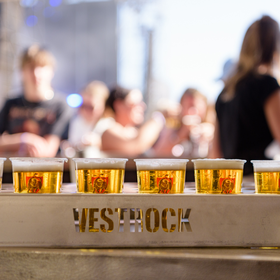 A wooden cup holder with the letters VESTROCK containing 4 beers against a backdrop of people